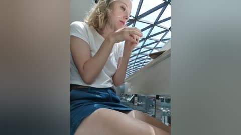 Media: A video of a blonde woman in a white t-shirt and denim skirt, sitting on a counter, holding a cup, in a modern room with a glass ceiling and stainless steel appliances.