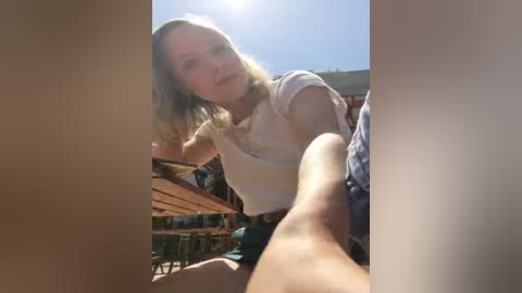 Media: Video of a blonde woman in a white t-shirt, sitting outdoors in sunlight, holding a beer bottle, with a blurred background of wooden tables and chairs.