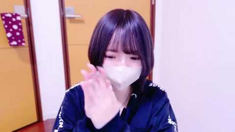 Media: Video of a young East Asian woman with straight, shoulder-length black hair, wearing a navy blue jacket, white face mask, and a white handkerchief covering her mouth, in a brightly lit room with a beige door and patterned cloth.