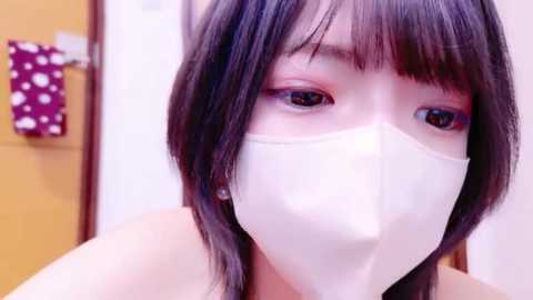 Media: Video of an Asian woman with straight black hair and bangs, wearing a white surgical mask, looking directly at the camera. Background includes a wooden door and a red polka-dotted cloth.