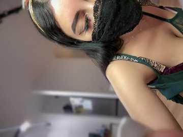 Media: Video of a young woman with long black hair, wearing a black mask, dark green spaghetti-strap top, and gold jewelry. She has a slim build and light skin tone, with her head tilted down and eyes closed. The background is blurred, showing a kitchen countertop.