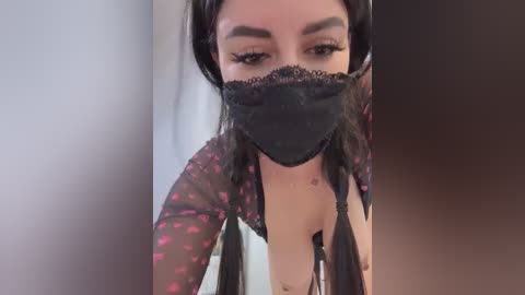 Media: Video of a woman with light brown skin, wearing a black lace mask, sheer pink floral robe, and black bra. She has long, dark hair and is indoors, looking down.