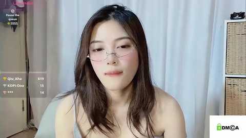 Media: Video of an East Asian woman with long dark hair, fair skin, and small glasses, smiling slightly, wearing a strapless top, in a modern indoor setting with a white curtain background.