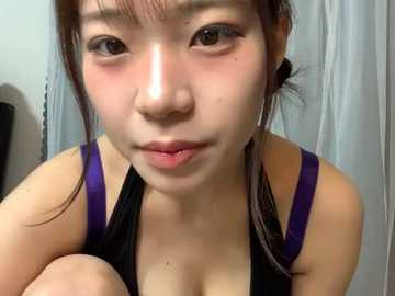 Media: Video of an East Asian woman with fair skin, brown eyes, and dark hair tied back, wearing a black tank top with purple straps, in a bathroom with white walls and a shower curtain.