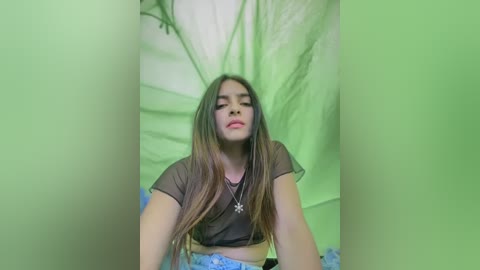 Media: A video of a young Latina woman with long brown hair, wearing a sheer black crop top and blue jeans, posing against a green fabric backdrop.