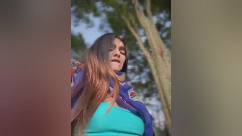 Media: Video of a young woman with long brown hair, wearing a blue shirt, standing outdoors with blurred green trees in the background, her expression serious.