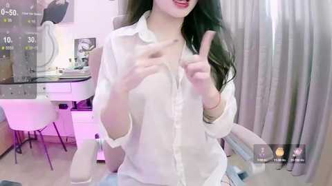 Media: Video of an East Asian woman with long, dark hair, wearing a sheer white blouse, sitting in a brightly lit room with modern furniture.