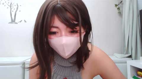 Media: Video of an Asian woman with straight brown hair, wearing a gray ribbed turtleneck and a pink face mask, in a white bathroom with minimalist decor.