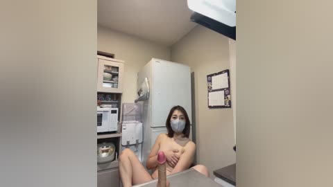 Media: Video of an Asian woman with shoulder-length brown hair, wearing a mask and nothing else, holding a large erect penis, in a small kitchen with white appliances and cabinets.