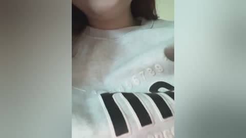 Media: Video of a person's neck and upper chest in a white, oversized graphic tee featuring black stripes and text, partially obscured by a blurred background.