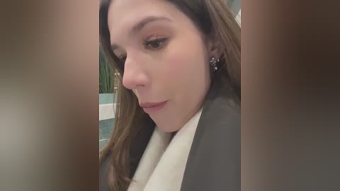 Media: Video of a young Caucasian woman with light skin, brown hair, and subtle makeup, wearing a white towel and a black robe, standing in a bathroom with tiled walls and a green shower curtain in the background.