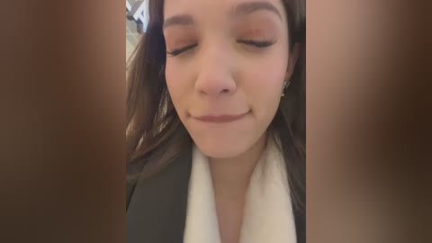 Media: Video of a young woman with light skin, closed eyes, and a slight smile, wearing a dark jacket over a white shirt, with her brown hair partially visible.