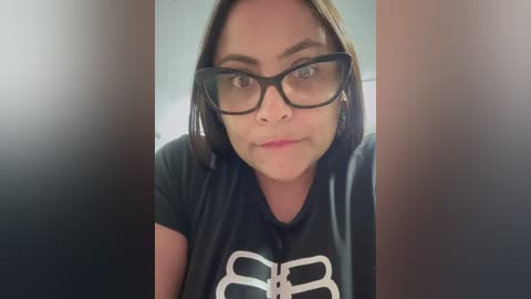 Media: Video of a woman with straight brown hair, wearing black-framed glasses and a black T-shirt with white design, indoors.