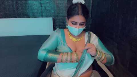 Media: Video of a South Asian woman in a teal saree, gold jewelry, and face mask, adjusting her blouse in a dimly lit room with dark walls.