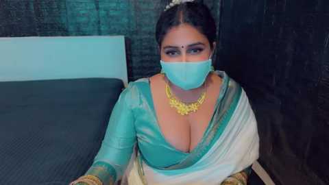 Media: Video of a woman in a teal saree and white shawl, wearing a gold necklace, turquoise face mask, and bindi, seated on a black bed against a dark wall.