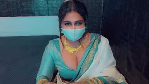 Media: Video of a South Asian woman in a teal sari with gold embroidery, wearing a face mask, gold necklace, and bindi, against a dark wall, showcasing traditional attire.