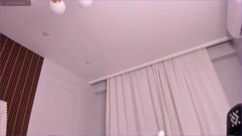 Media: Video of a minimalist, modern room with a white ceiling, beige curtains, brown striped wall, and a white wall. A circular light fixture hangs overhead.