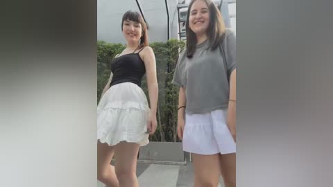 Media: Video of two smiling young women standing side-by-side outdoors, wearing casual summer outfits: one in a black top and white skirt, the other in a gray top and white shorts.