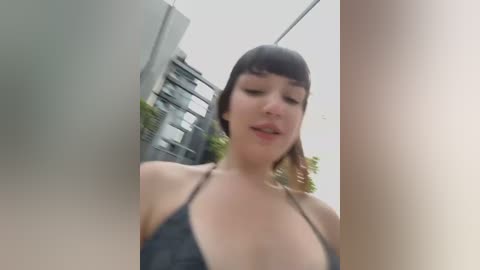 Media: A low-angle, blurred video of a young woman with short black hair, wearing a black bikini top, standing in an urban setting with modern buildings in the background.