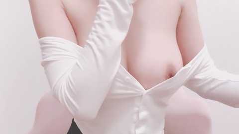 Media: Video of a fair-skinned, nude woman with medium-sized breasts, wearing a white, off-the-shoulder, button-up top, which is partially unbuttoned, revealing her left breast. The background is plain and light-colored, emphasizing the subject.