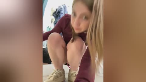 Media: Video of a young woman with long blonde hair, squatting in a room. She wears a maroon long-sleeve shirt and beige sneakers. Background shows a bed with blue and white bedding and a pile of clothes.