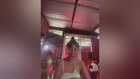 Media: A video of an Asian woman with medium skin tone, short black hair, and glasses, wearing a green bra and panties, standing in a dimly lit, rustic room with red curtains and a corrugated metal ceiling.