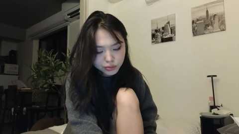 Media: Video of a young East Asian woman with long black hair, wearing a gray sweater, sitting on a bed in a dimly lit room with black-and-white framed photos on the wall.