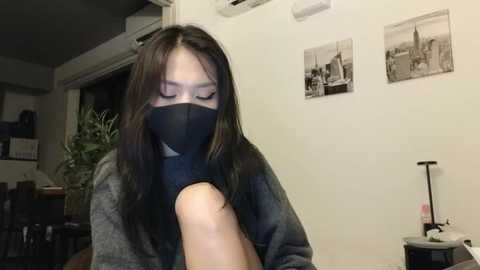 Media: A video of a person with long black hair, wearing a black face mask and gray sweater, holding a white object, in a dimly lit room with framed black-and-white photos on the wall.