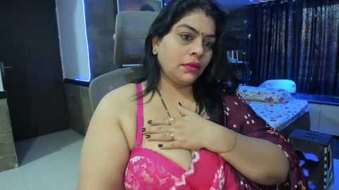 Media: A video of a plus-sized South Asian woman with long black hair, wearing a pink floral-patterned bra, sitting in a dimly lit room with a bed and window in the background.