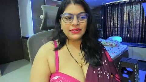 Media: Video of a middle-aged South Asian woman with medium brown skin and long black hair, wearing glasses and a pink lace bra, sitting in a modern bedroom with dark curtains and a bed.