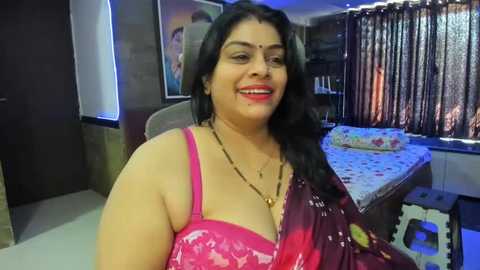 Media: Video of a smiling, curvy South Asian woman with long black hair, wearing a pink bra with a floral pattern, and a maroon sari, in a dimly lit bedroom with a bed and floral curtains.