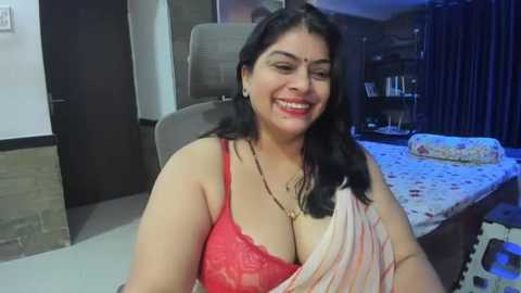 Media: Video of a smiling South Asian woman with medium skin tone, dark hair, and wearing a red lace bra and a pink sari. She is indoors, in a modern bedroom with a bed, dark curtains, and a soft carpet.