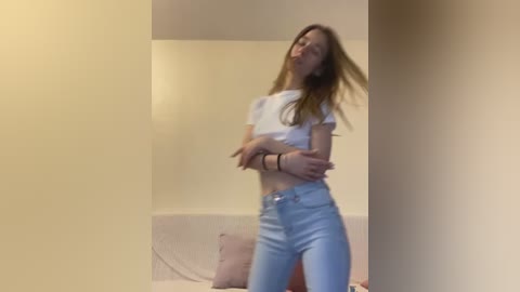 Media: Video of a young, slender, Caucasian woman with long, light brown hair, wearing a white cropped top and light blue jeans, posing in a small, beige-walled room.