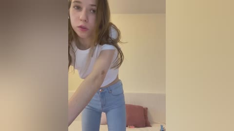 Media: Video of a young woman with light skin and long brown hair, wearing a white crop top and high-waisted blue jeans, bending over a table in a minimalist, beige-colored room.