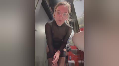 Media: Video of a slender, fair-skinned woman with blonde hair in a ponytail, wearing a sheer black bodysuit and glasses, bending over in a dimly lit, cluttered room with industrial elements.