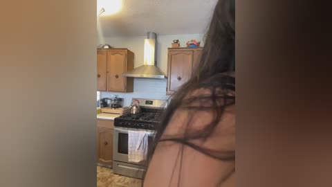 Media: Video of a small, cozy kitchen with wooden cabinets, a stainless steel range, and a white towel hanging over the oven. A woman with long brown hair stands partially obscured in the foreground, her back to the camera.