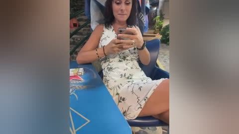 Media: Video of a woman in a white floral dress, seated on a blue inflatable lounge chair, taking a selfie with a smartphone.