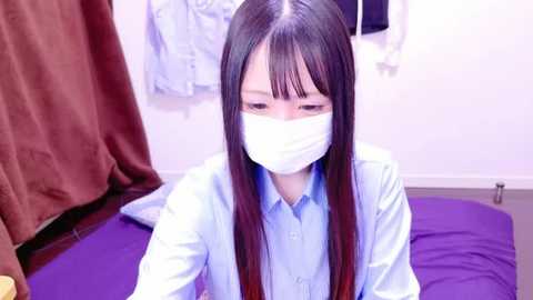 Media: Video of an Asian woman with long straight black hair and fair skin, wearing a light blue shirt and white mask, seated on a purple bed in a room with a white wall and brown curtain.