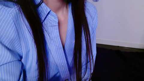 Media: A video of a woman with long, straight dark brown hair, wearing a light blue, vertically striped button-up shirt, revealing cleavage, against a white wall and dark carpeted floor.