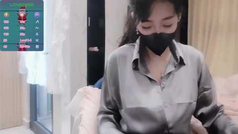 Media: Video of an East Asian woman with long black hair, wearing a gray shirt and black mask, seated in a room with a wooden door and white curtains.