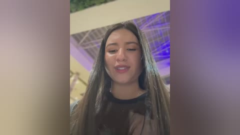 Media: Video of a young woman with long, straight brown hair, wearing a dark top, smiling, standing indoors with a purple and white geometric ceiling in the background.