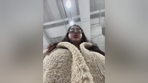 Media: Video of a young woman with long brown hair and glasses, wearing a fluffy beige cardigan, standing in a dimly lit industrial space with exposed white beams and pipes.