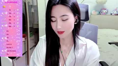 Media: Video of an East Asian woman with long black hair, fair skin, wearing a white shirt, red lipstick, sitting in a room with a bed, toys, and a pink screen.