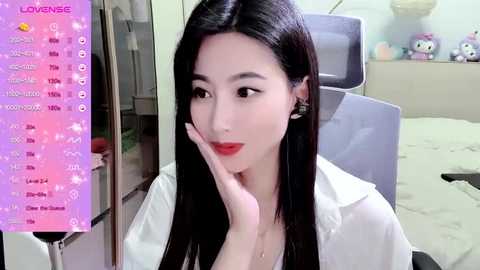 Media: Video of a young East Asian woman with long black hair, wearing a white lab coat, looking contemplative, in a modern bedroom with a bed and stuffed animals.