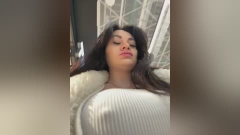 Media: A video of a woman with long, dark hair and light skin, wearing a white ribbed sweater, lying on a white fur blanket. The background features a white, geometric window frame.