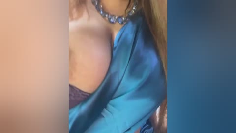 Media: Video of a woman with light skin and curly brown hair, wearing a blue satin robe, revealing her cleavage and a lacy bra. She has a chunky silver necklace. The background is blurred.