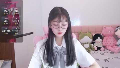 Media: Video of an East Asian woman with long black hair, wearing glasses, a white shirt, and a grey bow tie, sitting in a pink gaming chair in a cozy, stuffed animal-filled room.