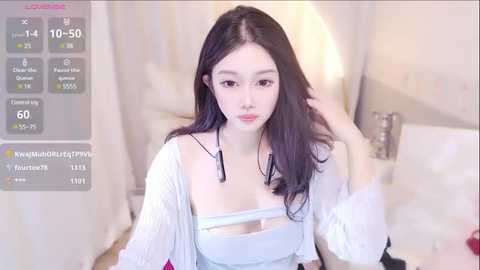 Media: Video of a young East Asian woman with long black hair, fair skin, and small breasts, wearing a white lace top and a sheer cardigan, in a softly lit bedroom.