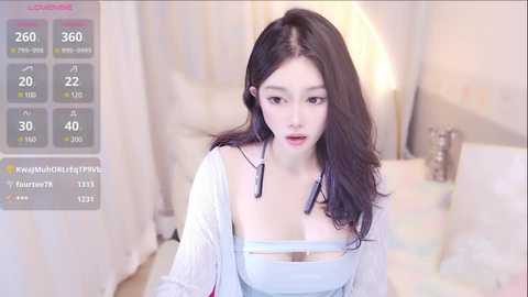 Media: Video of an East Asian woman with long black hair, fair skin, and wearing a white crop top, standing in a softly lit bedroom with a bed and lamp.