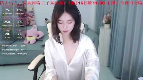 Media: Video of an Asian woman with shoulder-length black hair, wearing a white silk shirt, sitting on a light grey chair in a modern living room. Background features a pink plush toy and a plant.